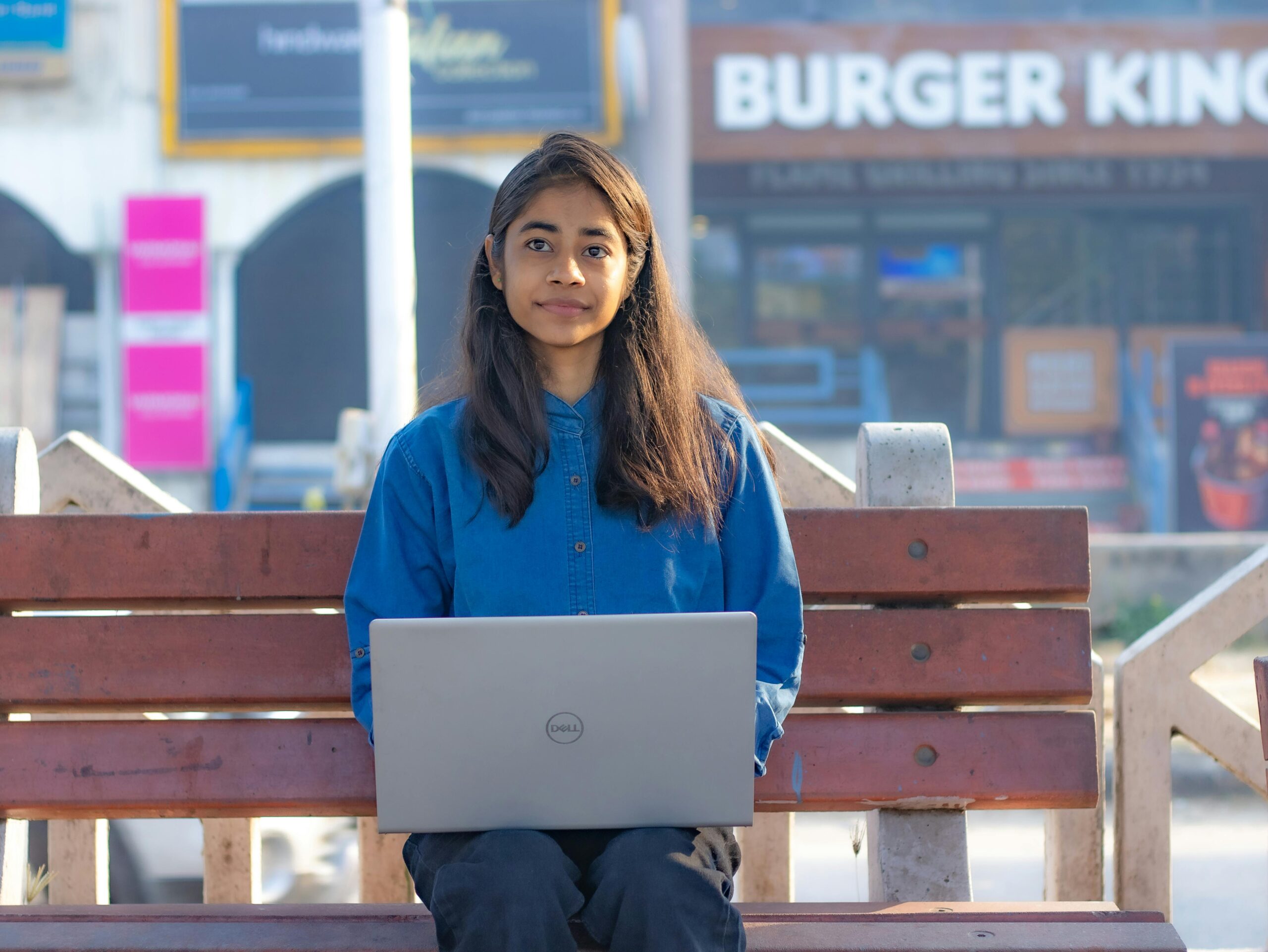 Teenage girl sitting on a bench using a laptop in an urban outdoor setting.