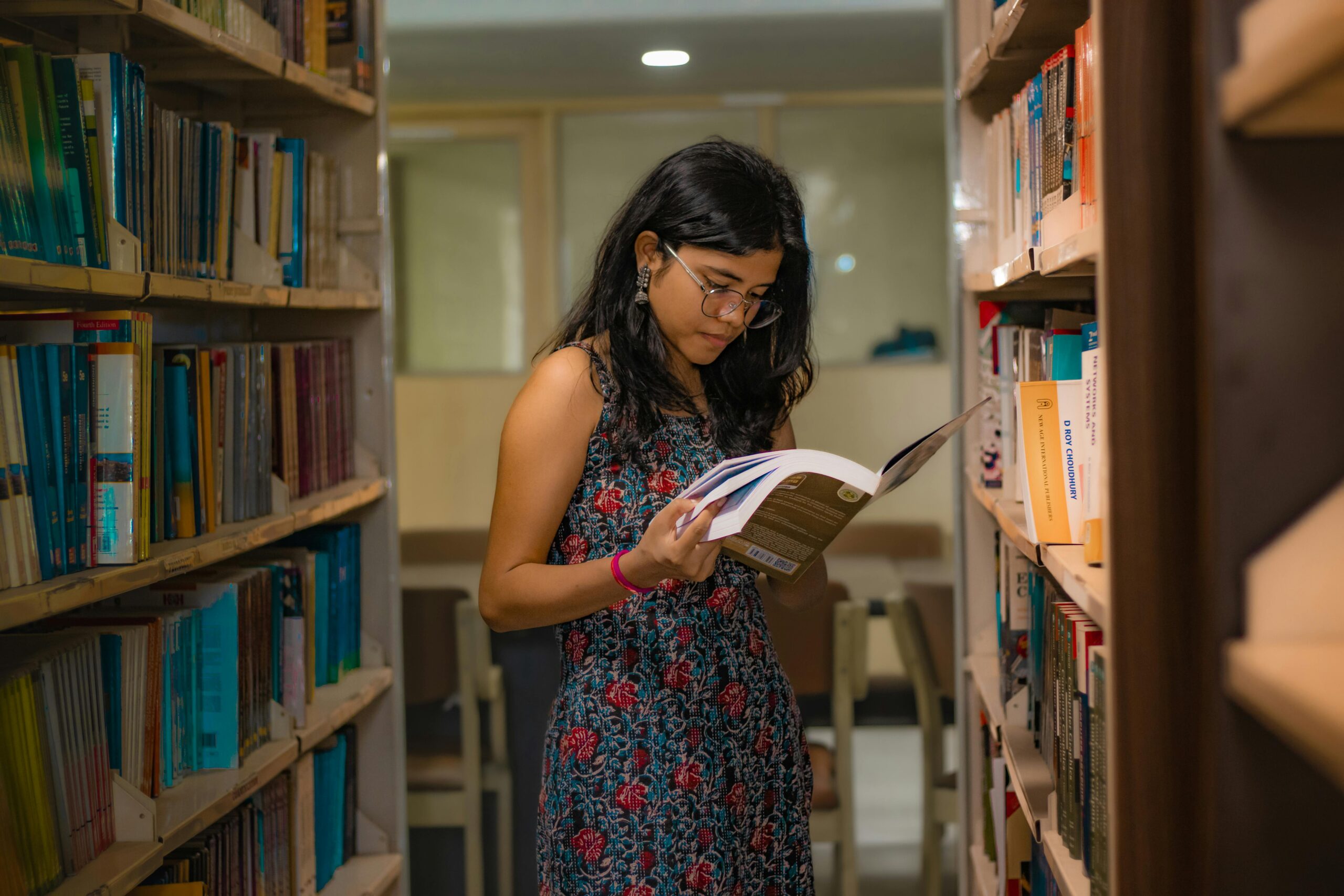 A South Asian woman with glasses reads attentively in a Delhi library, surrounded by bookshelves.